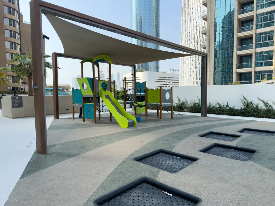 Urban rooftop playground with a bright green slide and climbing structures beneath a shade canopy, surrounded by tall buildings.
