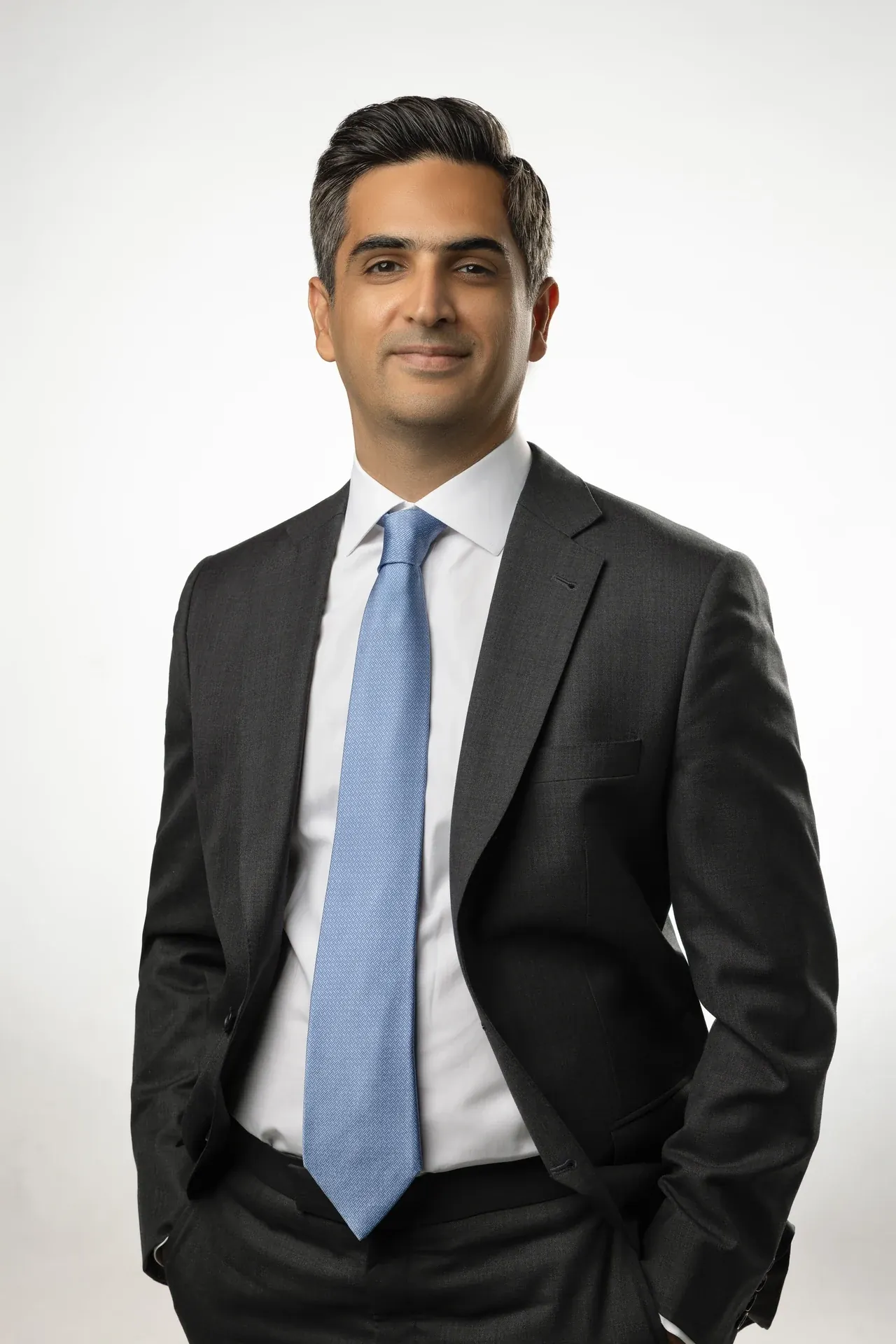 Professional man in a dark suit and light blue tie, standing with hands in pockets against a light background