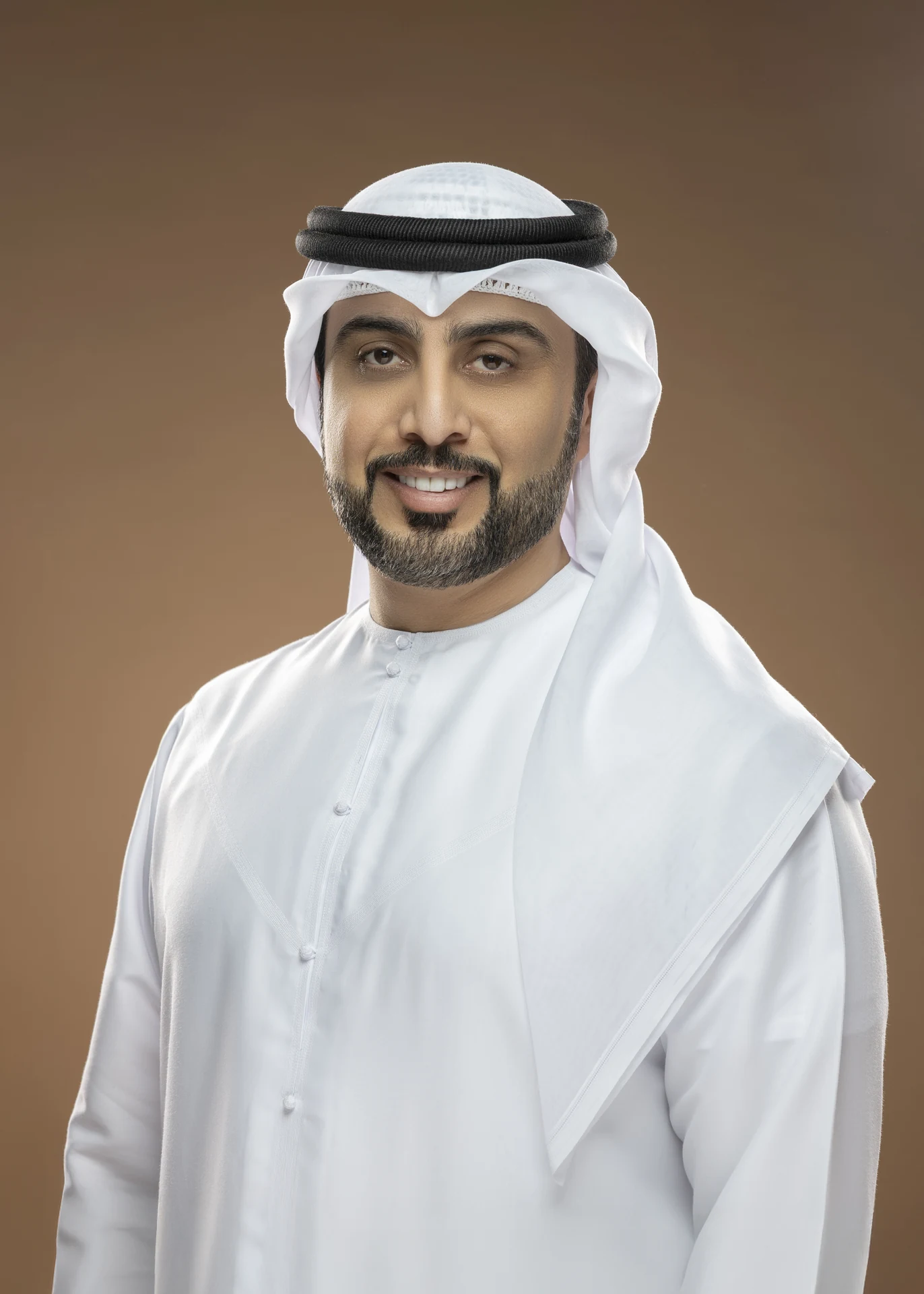 Portrait of a smiling man in traditional Emirati attire: white kandura and ghutra with agal, posing against a brown studio backdrop.