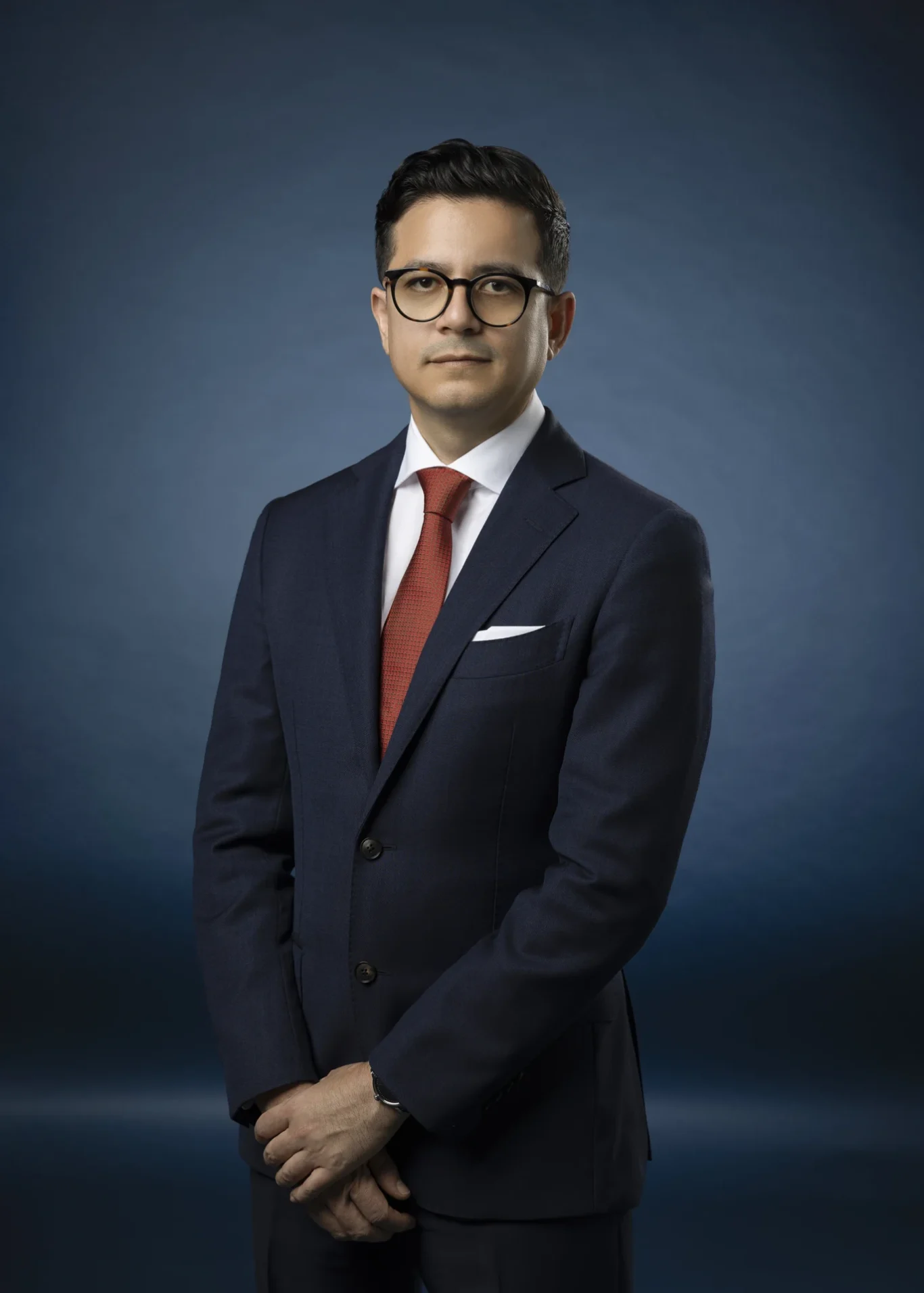Professional portrait of a man in a navy suit, white shirt, red tie, and round glasses against a blue gradient backdrop.