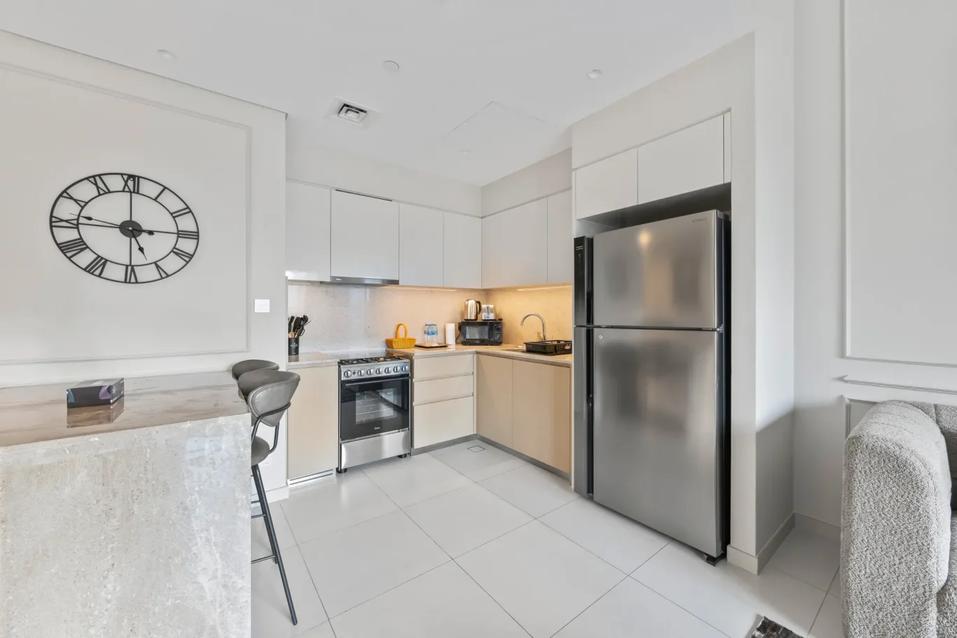 Modern kitchen with white cabinets, stainless steel fridge, and a large black wall clock on the left wall.