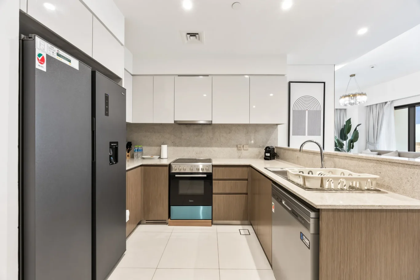 Modern kitchen with stainless steel fridge on the left, white upper cabinets, and a beige marble countertop island with a sink and dish rack in the foreground.