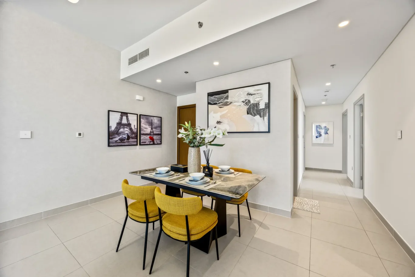 Modern dining area with a marble-top table set for four, yellow upholstered chairs, and framed artwork on light-gray walls.