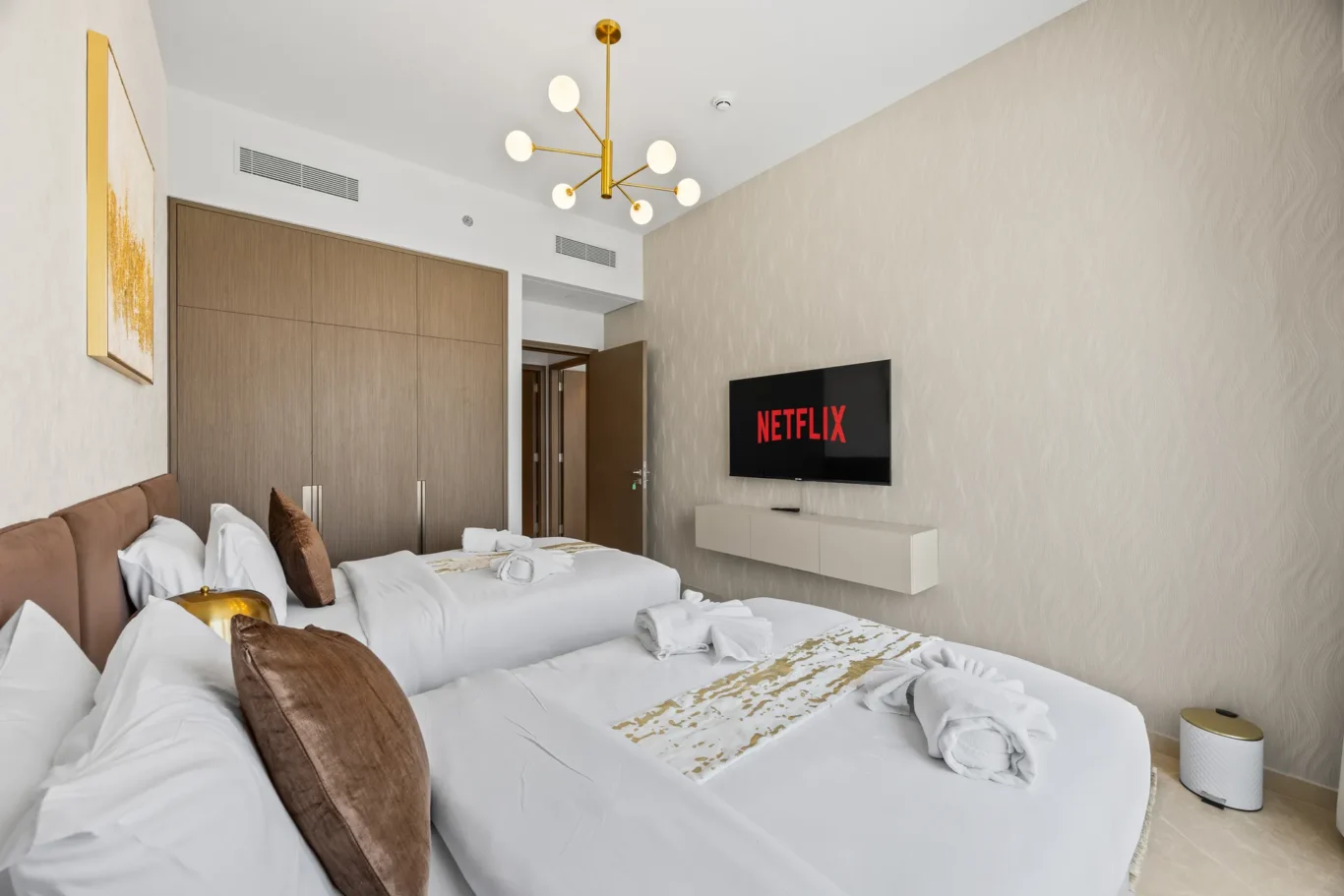 Two white-bedding hotel beds with brown cushions in a modern room; a wall-mounted Netflix TV and a gold chandelier above.