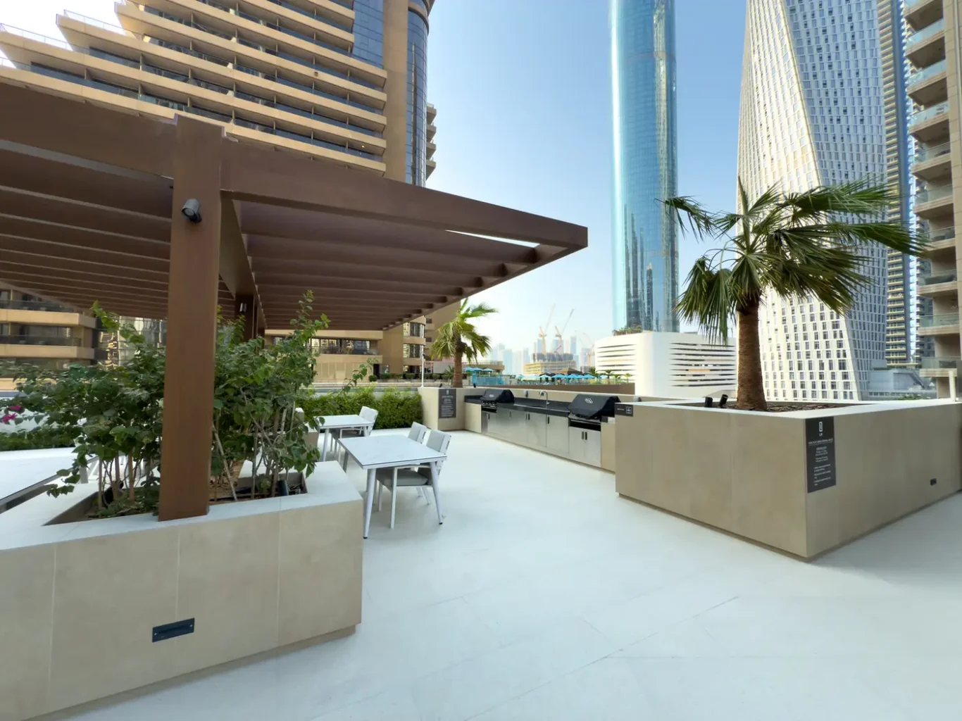 Rooftop BBQ area with built-in grills, seating, and planter boxes against a backdrop of tall glass skyscrapers.