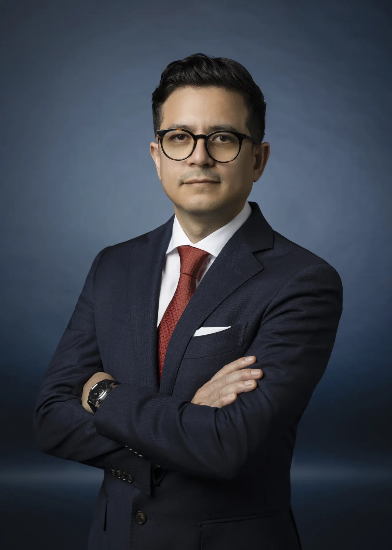 Professional man in a navy suit with red tie and glasses, arms crossed against a blue gradient backdrop.