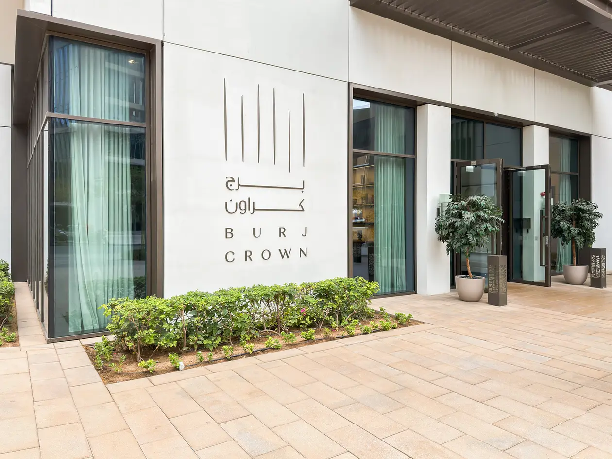 Exterior of a modern building entrance with large glass doors, beige tiles, and a sign reading 'Burj Crown' in English and Arabic, flanked by potted plants.