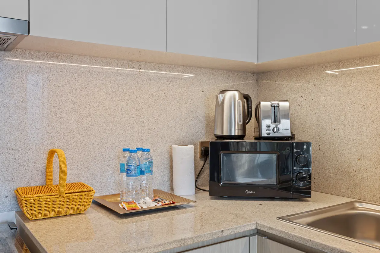 Kitchen counter with a black microwave, silver kettle, and toaster atop; water bottles, paper towels, and a wicker basket nearby.
