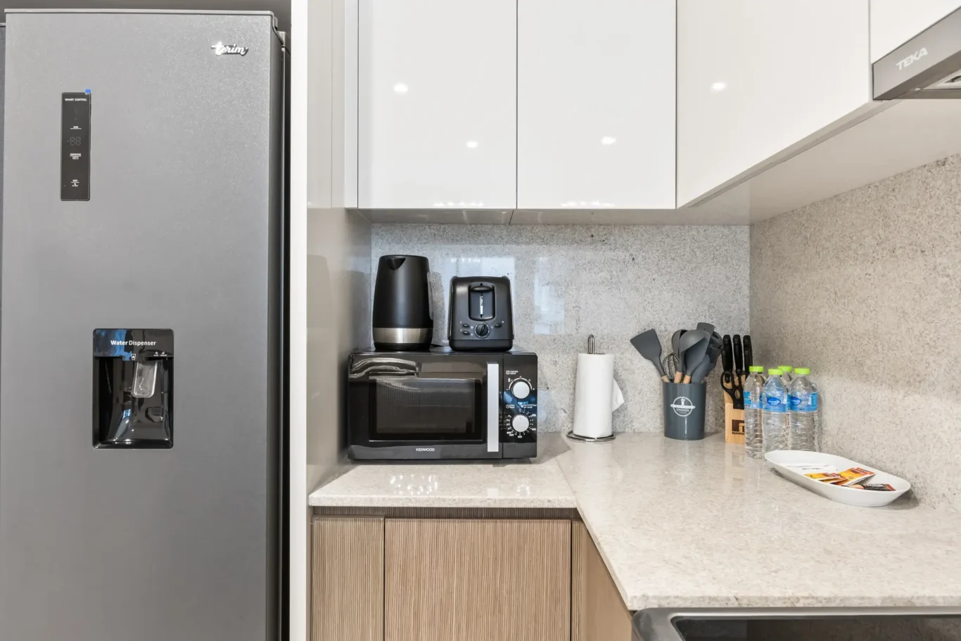 Tall gray refrigerator on the left with water dispenser, white glossy upper cabinets, and small appliances (kettle, toaster) on a speckled countertop.