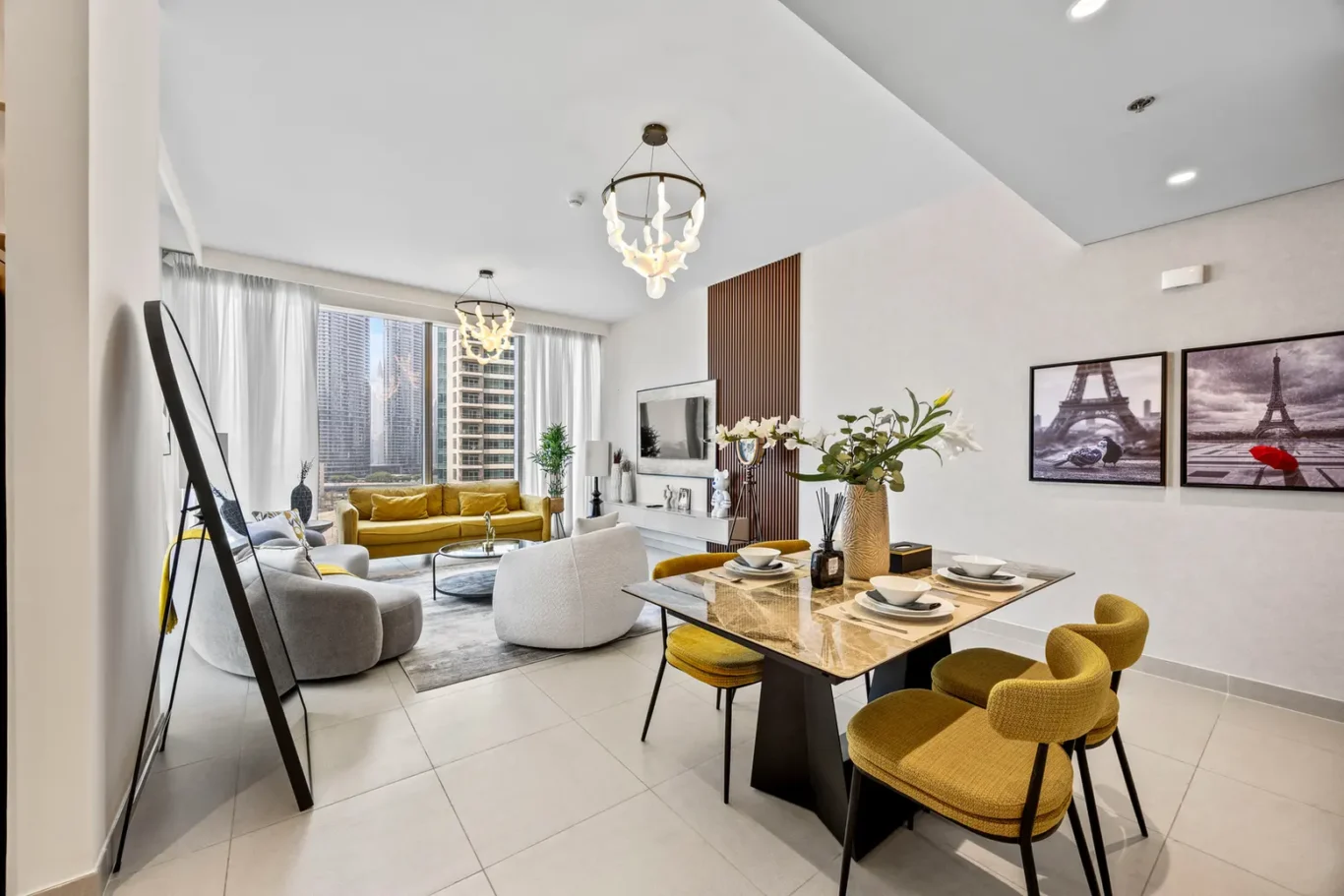 Open-concept living and dining room with a mustard-yellow sofa, grey lounge chairs, and a marble dining table by large city-view windows; neutral decor and plants add warmth.