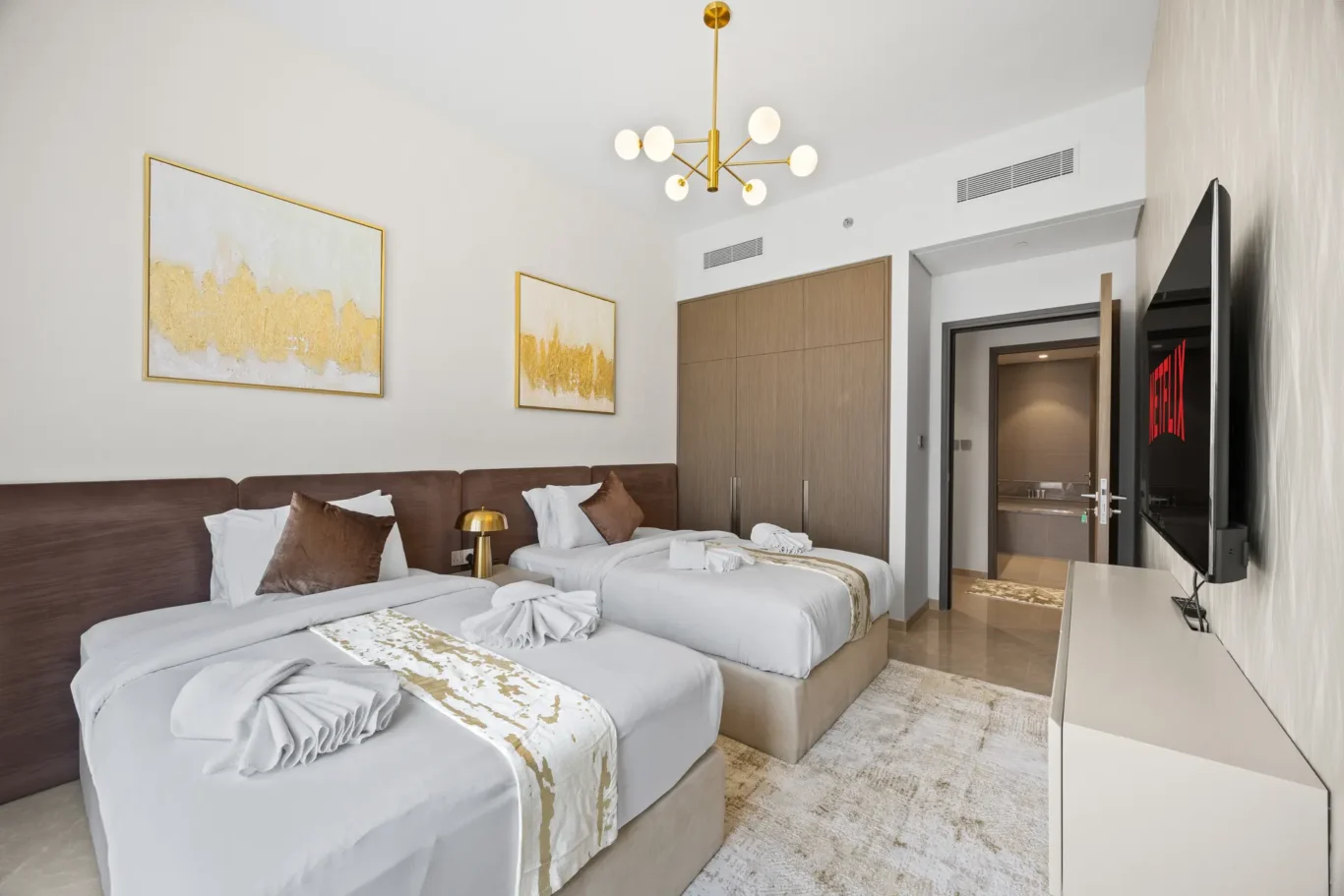 Hotel room with two twin beds, white linens, gold accents, and a wall-mounted flat-screen TV.