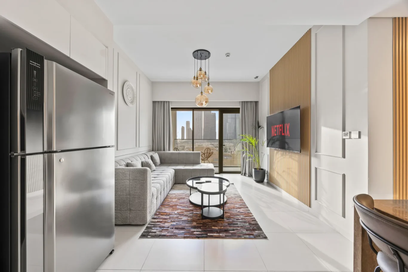 Modern open-plan living room with stainless steel fridge on the left, gray sectional sofa, round coffee tables, and a Netflix screen on the wall.