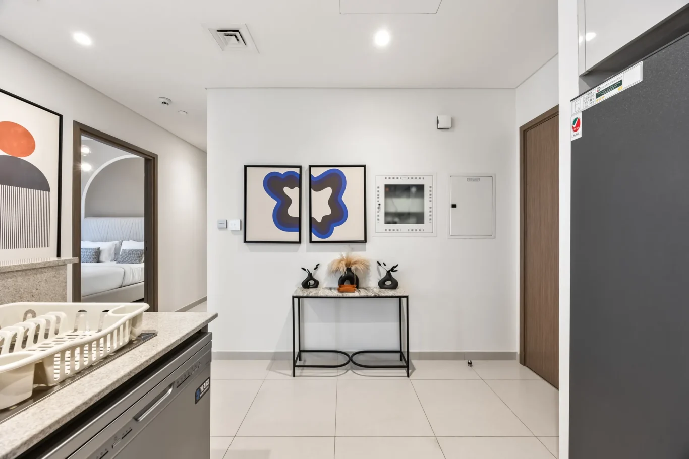 Modern kitchen with a dish rack on a granite counter and a dishwasher below; doorway to a bedroom in the background.