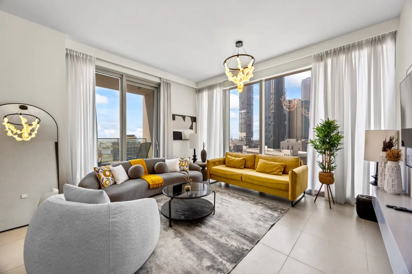 Bright, modern living room with large windows, a gray sectional and a mustard-yellow sofa, round glass coffee table, and a neutral area rug.