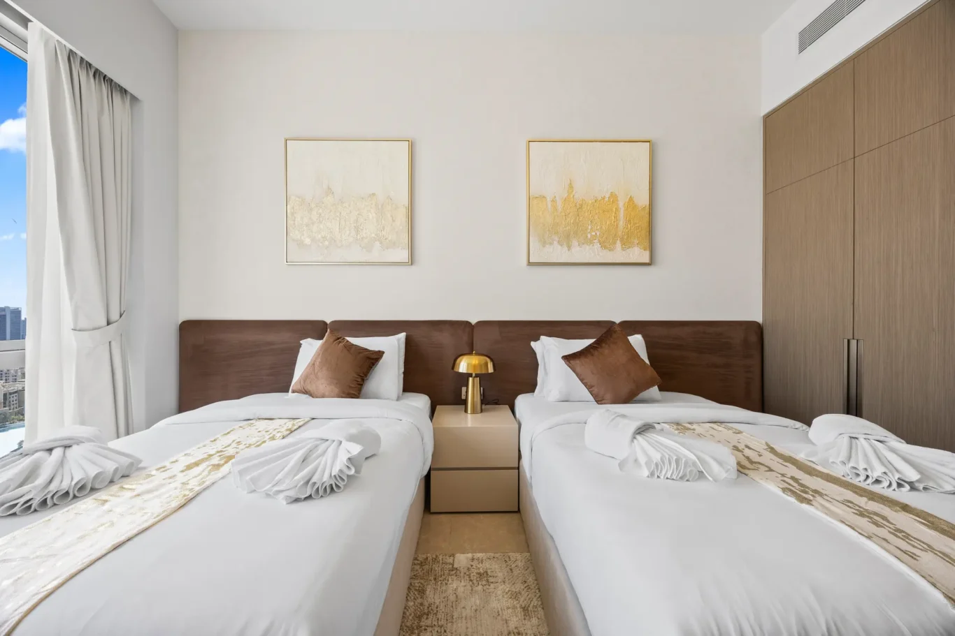 Twin hotel room with two single beds, white linens, brown pillows, and a shared headboard between a beige nightstand and gold lamp.