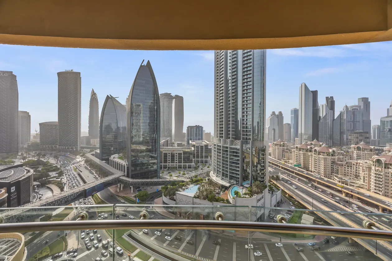 City skyline with modern skyscrapers and a busy multi-lane road, viewed from a balcony with a glass railing.