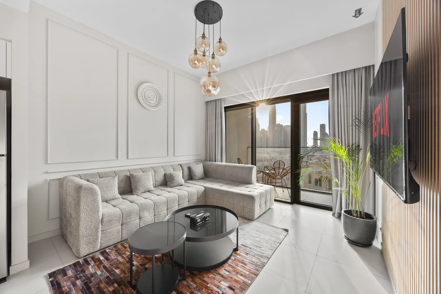 Modern living room with a beige sectional sofa, round black coffee tables, and a wall-mounted Netflix TV near a balcony with a city view.
