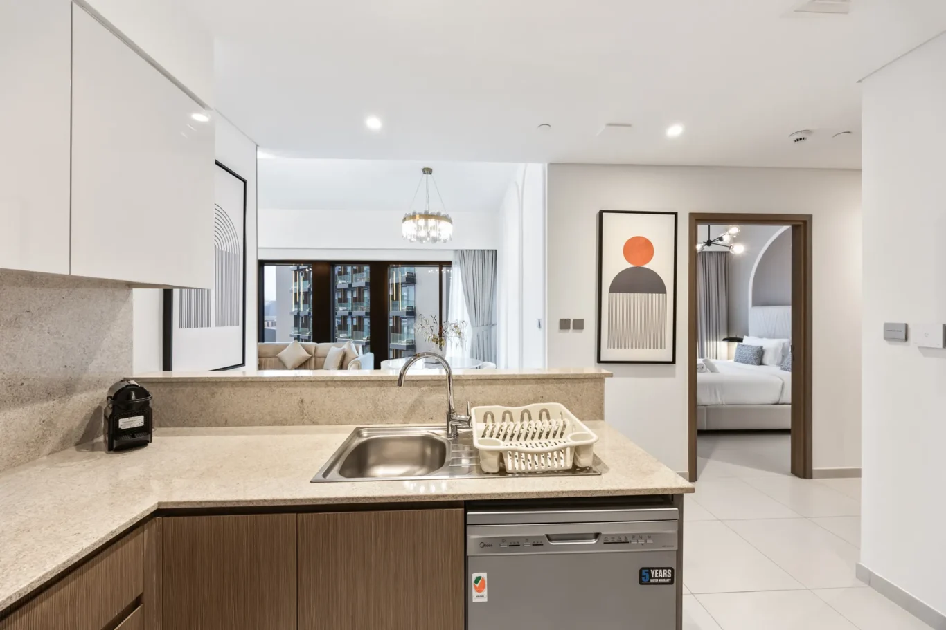 Open-plan kitchen with beige countertop, sink and dish rack; living room and bedroom visible beyond.
