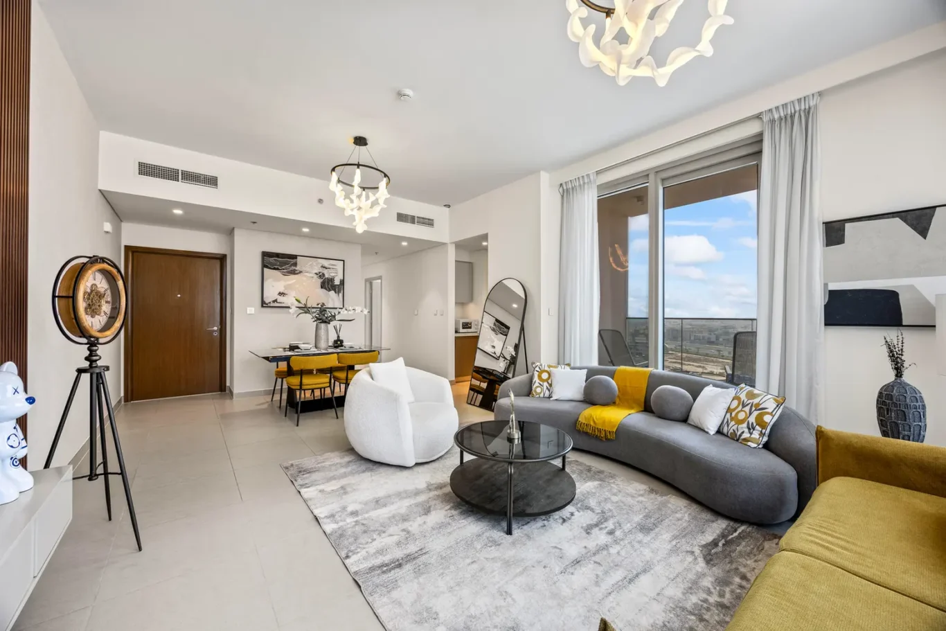 Open-concept living room with a curved gray sofa, yellow cushions, and a glass coffee table near large windows.