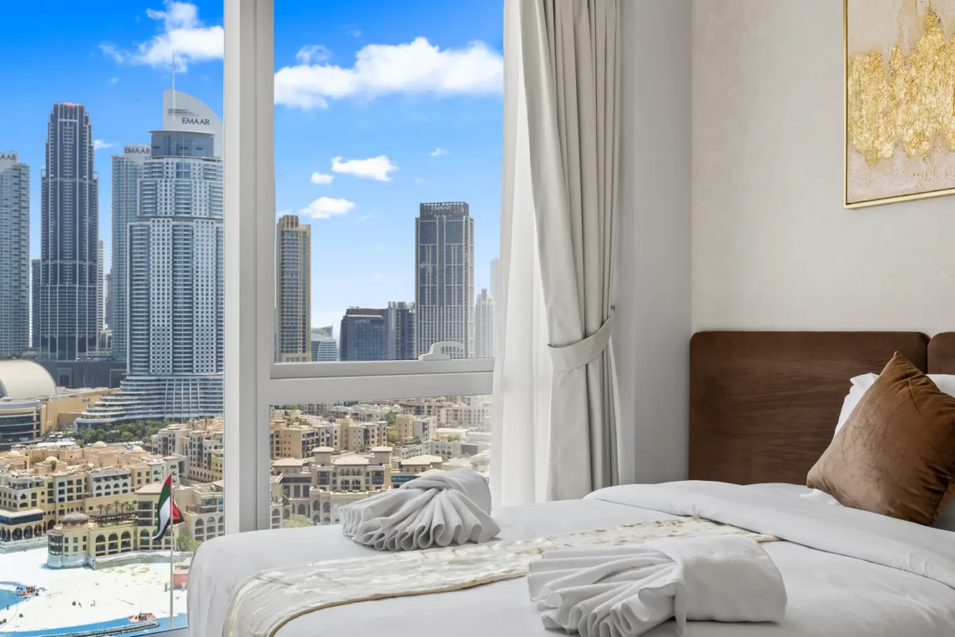 Luxurious hotel bedroom with white bedding and brown headboard, folded towel art on the bed, and a city skyline outside the window.