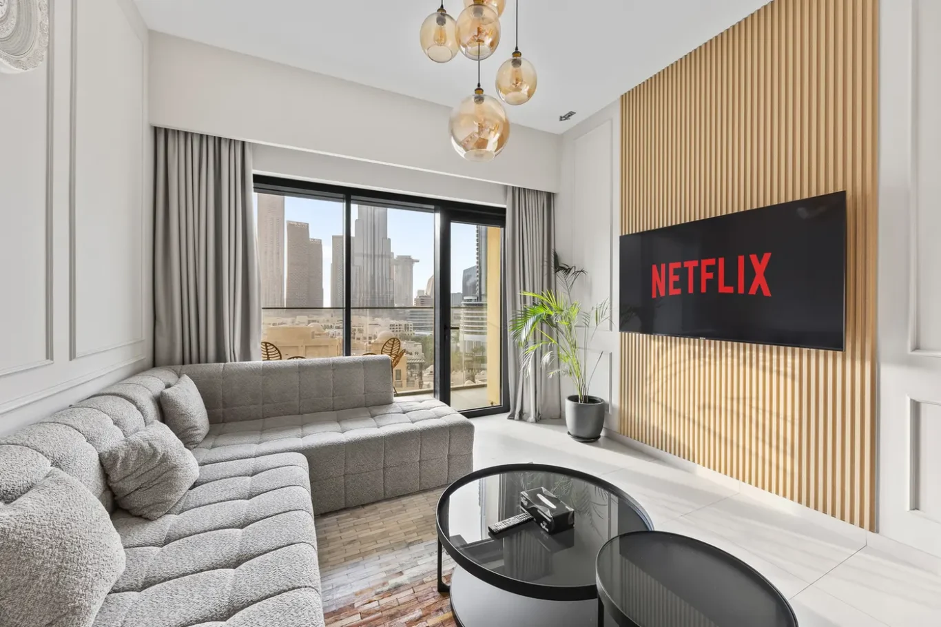 Modern living room with a gray sectional, glass coffee tables, plant, and a Netflix screen on a wood-slat wall.