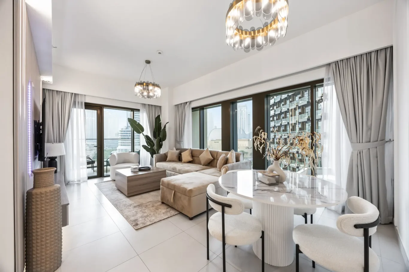 Bright living room with beige sectional, round marble dining table, and large windows showing city buildings; modern chandelier lighting the space.