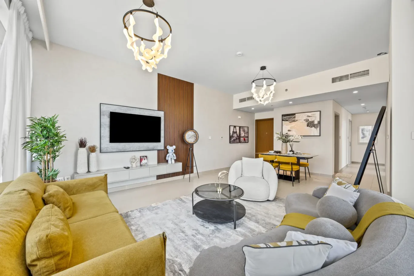 Bright open-concept living room with a mustard-yellow sofa, gray curved sofa, and glass coffee table, plus a dining area in the background.