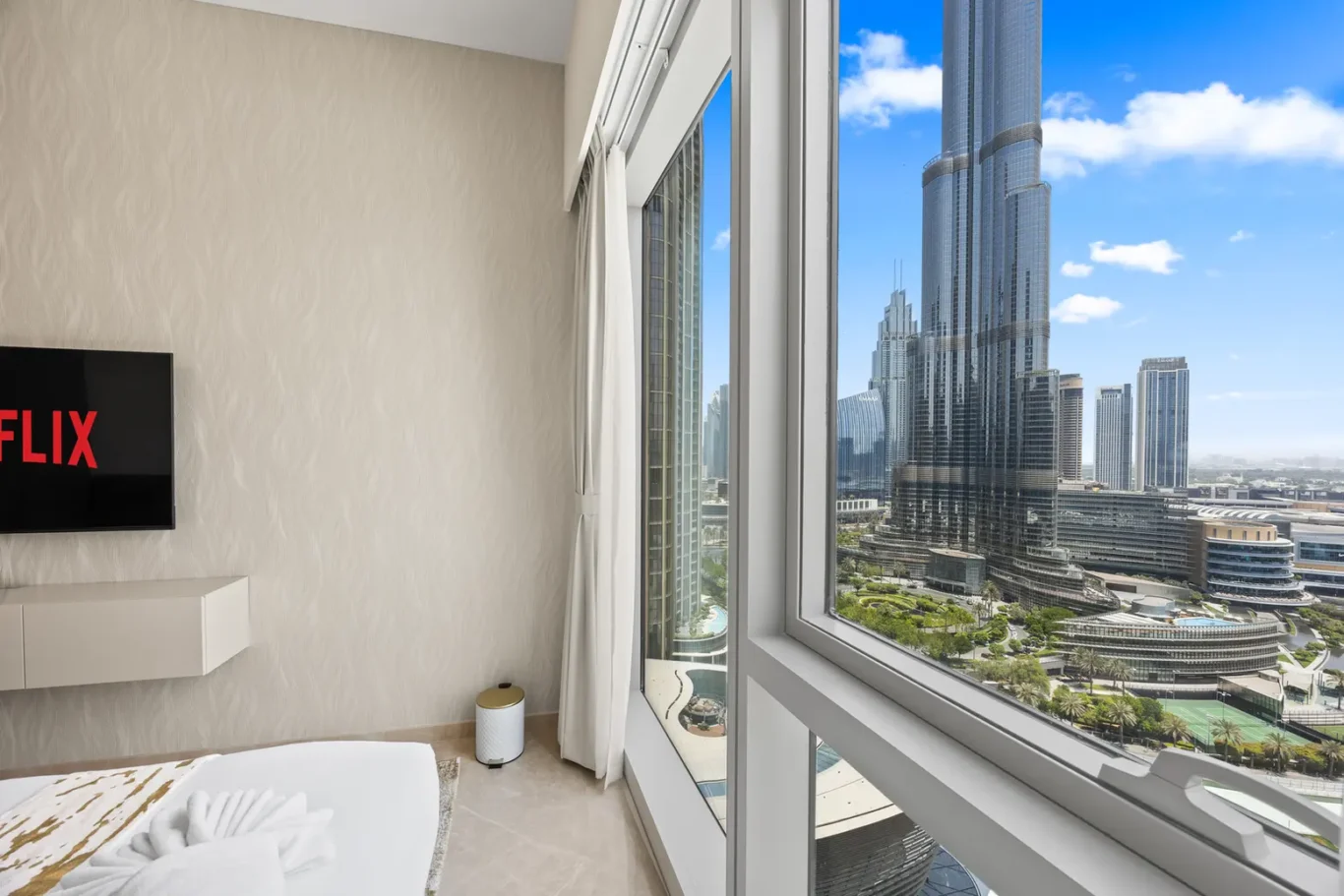 Hotel room with a large window offering a city view of modern skyscrapers and a pool area below; a flat-screen Netflix TV is mounted on a textured wall with a white shelf below.