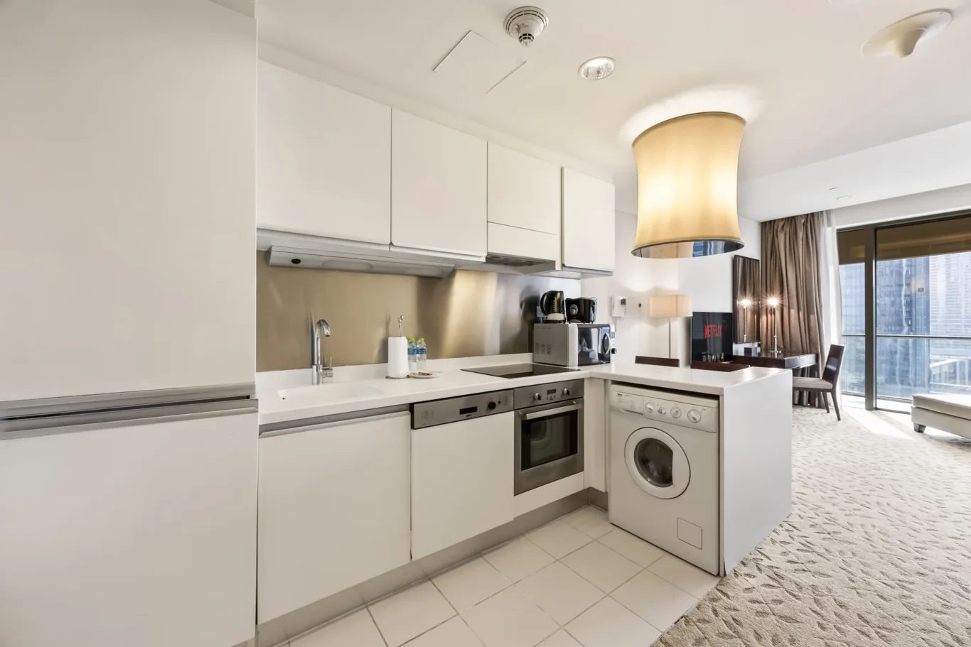 Modern white kitchen with stainless appliances, sink, and coffee station in a high-rise apartment living area.