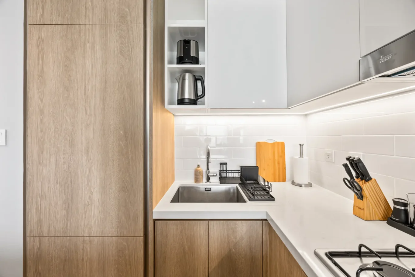 Modern kitchen countertop with white surface, wooden lower cabinets, and a stainless steel sink; dish rack and knife block beside the sink.