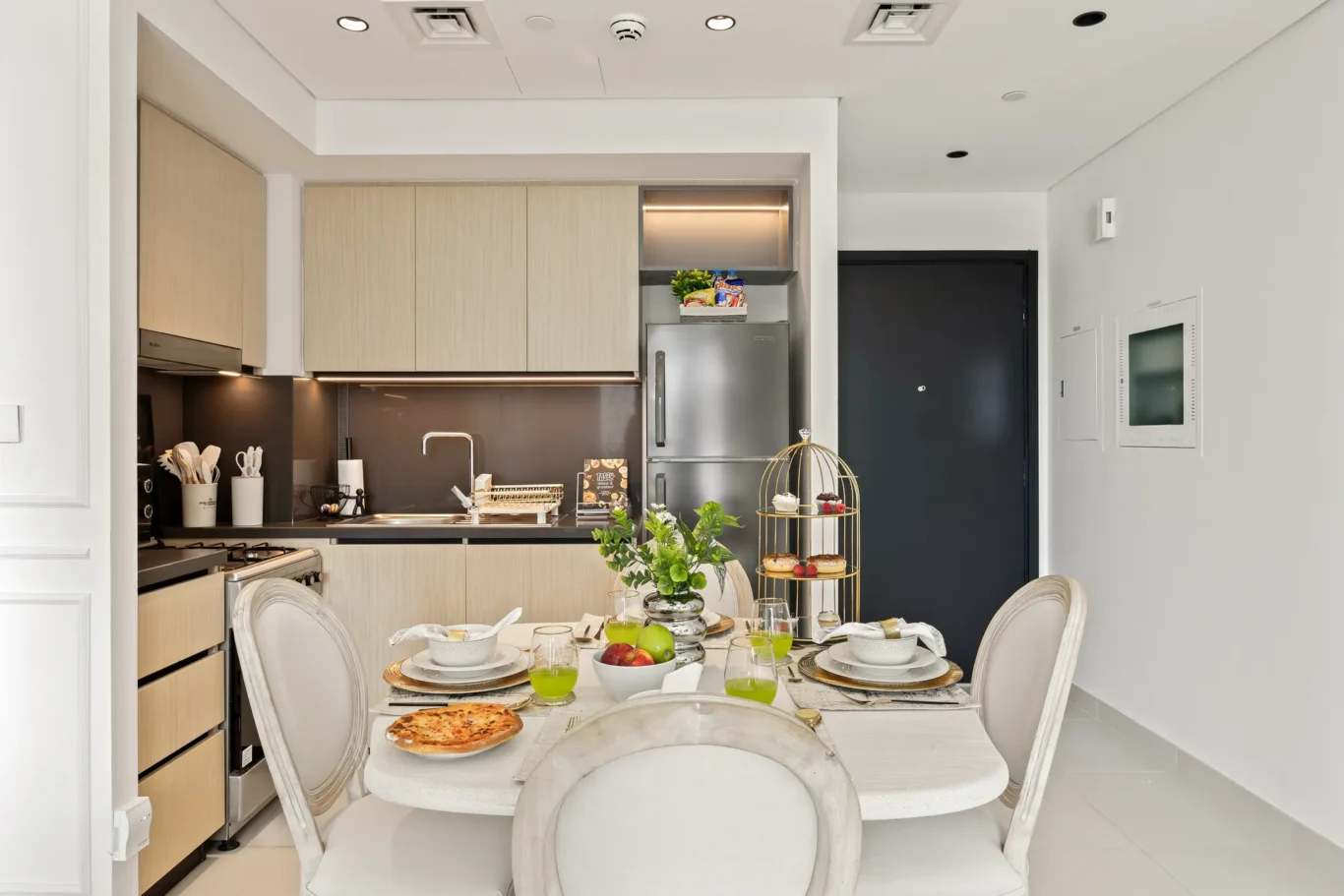 Modern kitchen and dining area with light wood cabinets, stainless fridge, and a tiered dessert stand on a dining table set for a meal.