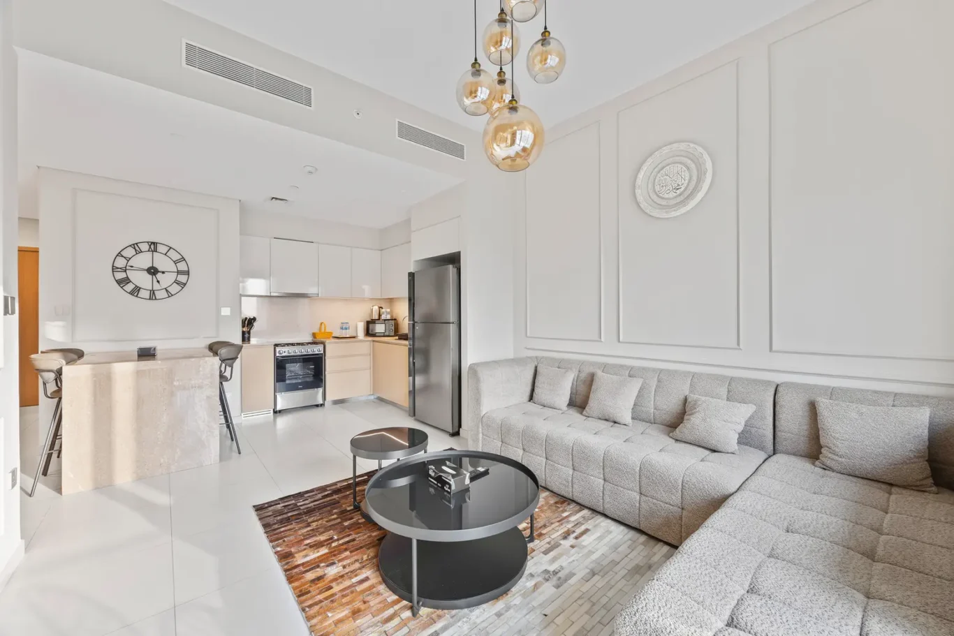 Open-plan living area with a light gray sectional sofa, glass coffee tables, and a beige kitchen island with stools against a white wall.