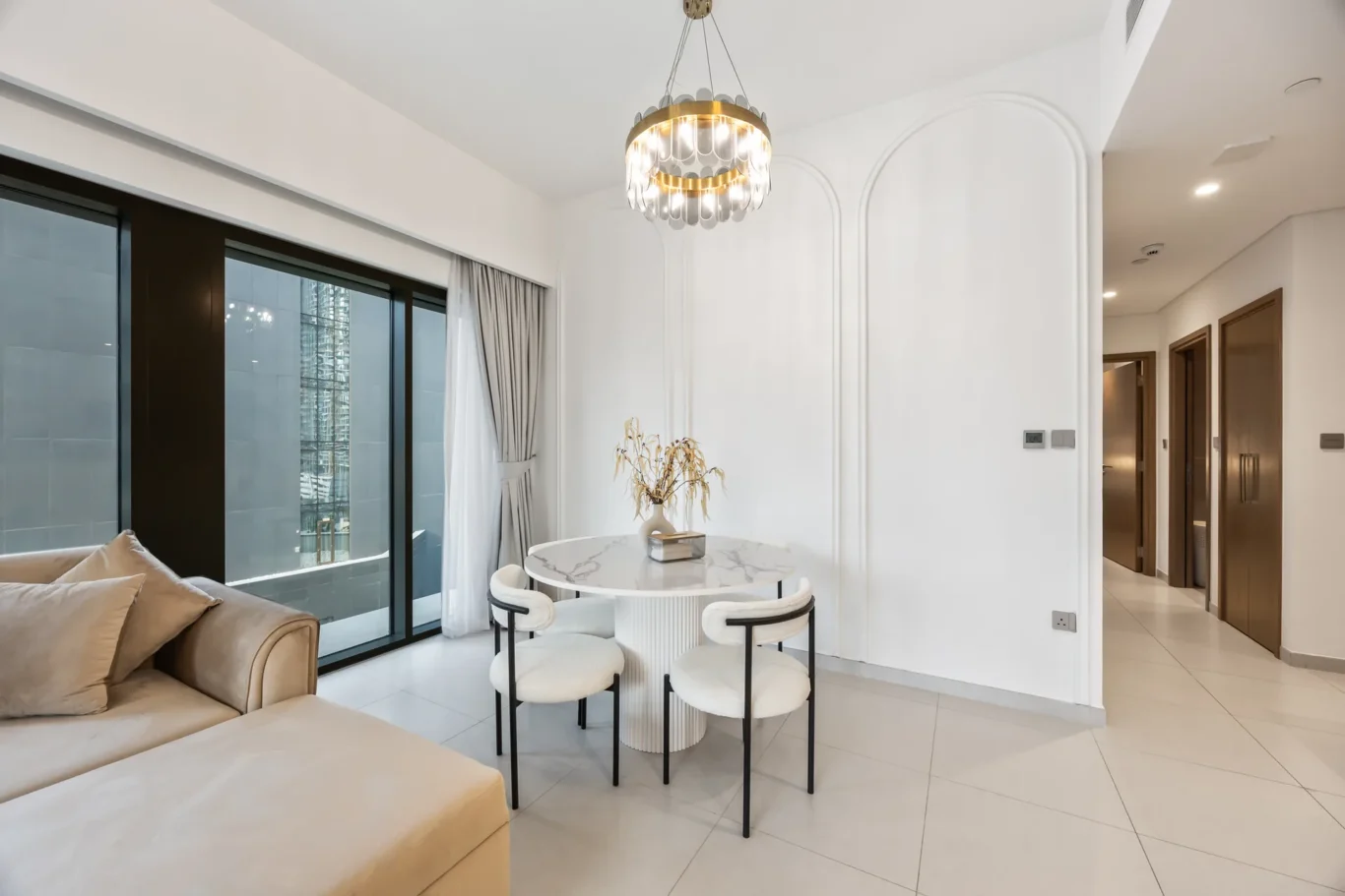 Bright living and dining area with a round marble table, four white stools, beige sofa, and a gold chandelier.