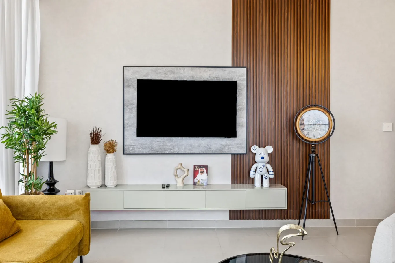 Living room with a wall-mounted TV in a gray framed surround, wooden slat panel to the right, and decor on a white console including a clock on a tripod.