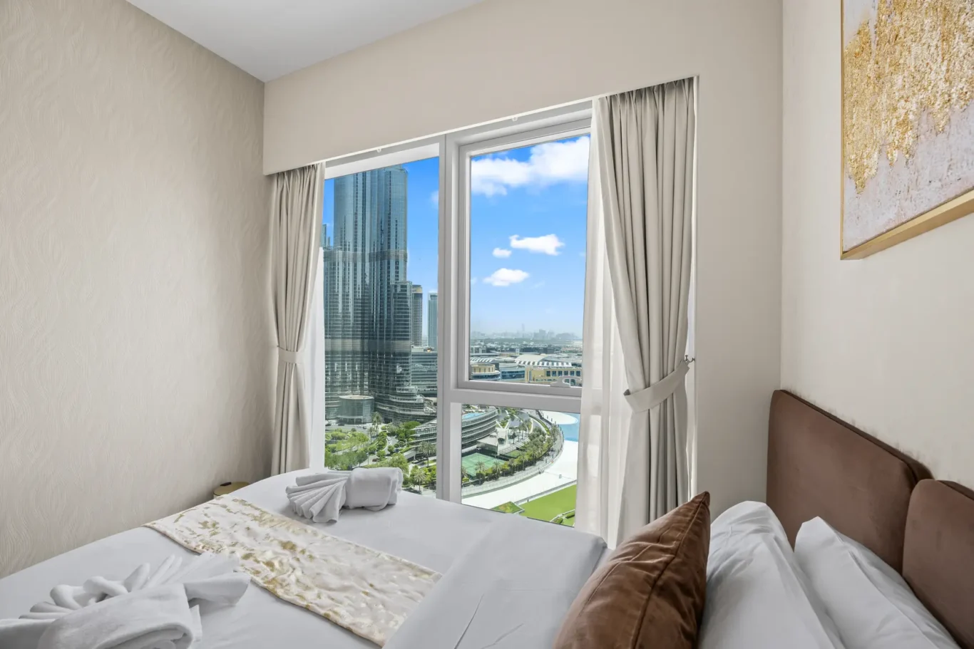 Hotel room view with a large window opening to a cityscape of modern skyscrapers and a curved waterfront, white bedding with brown cushions, and beige walls.