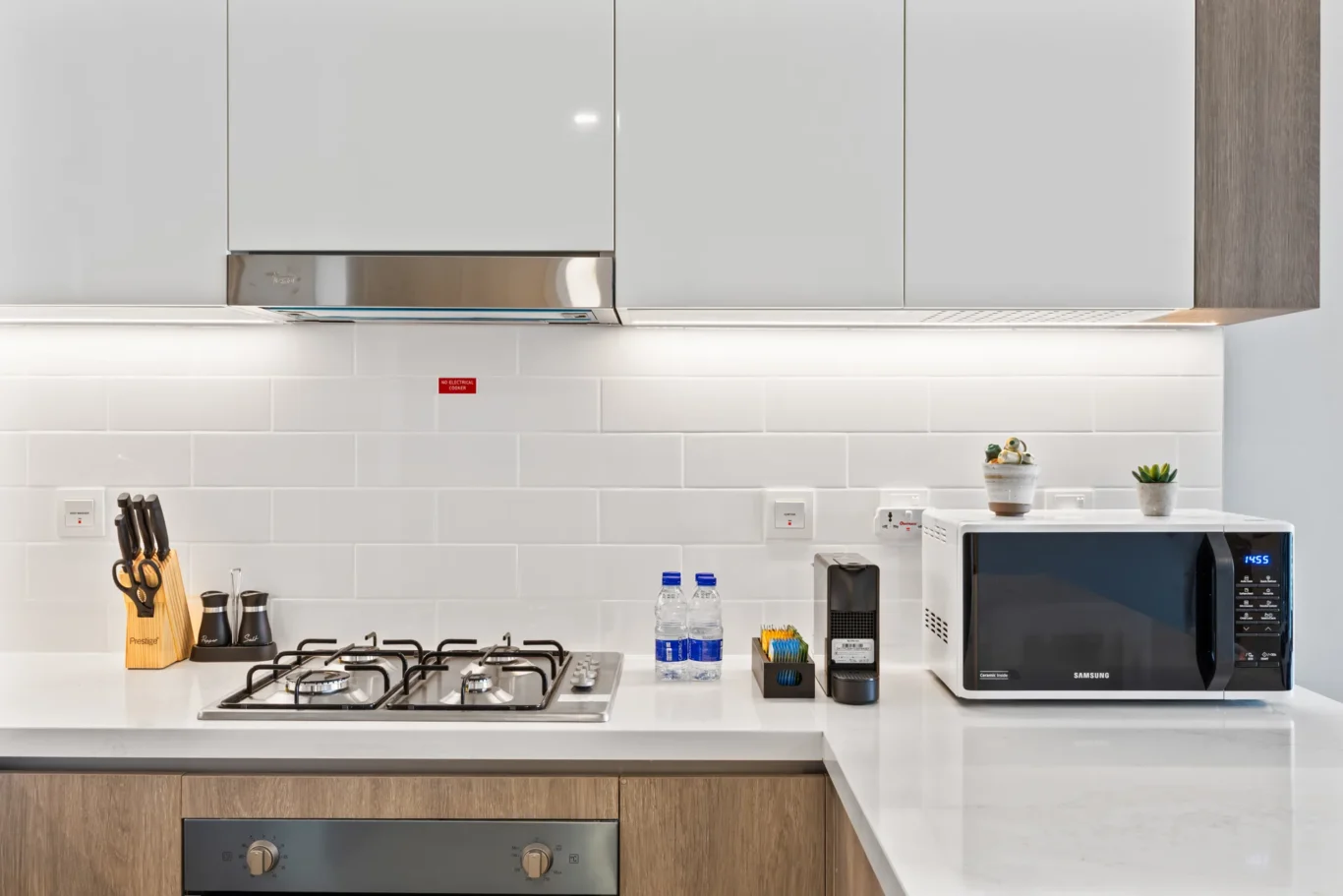 Modern kitchen scene with a gas cooktop, white countertops, and white upper cabinets; microwave and small plants on the counter.