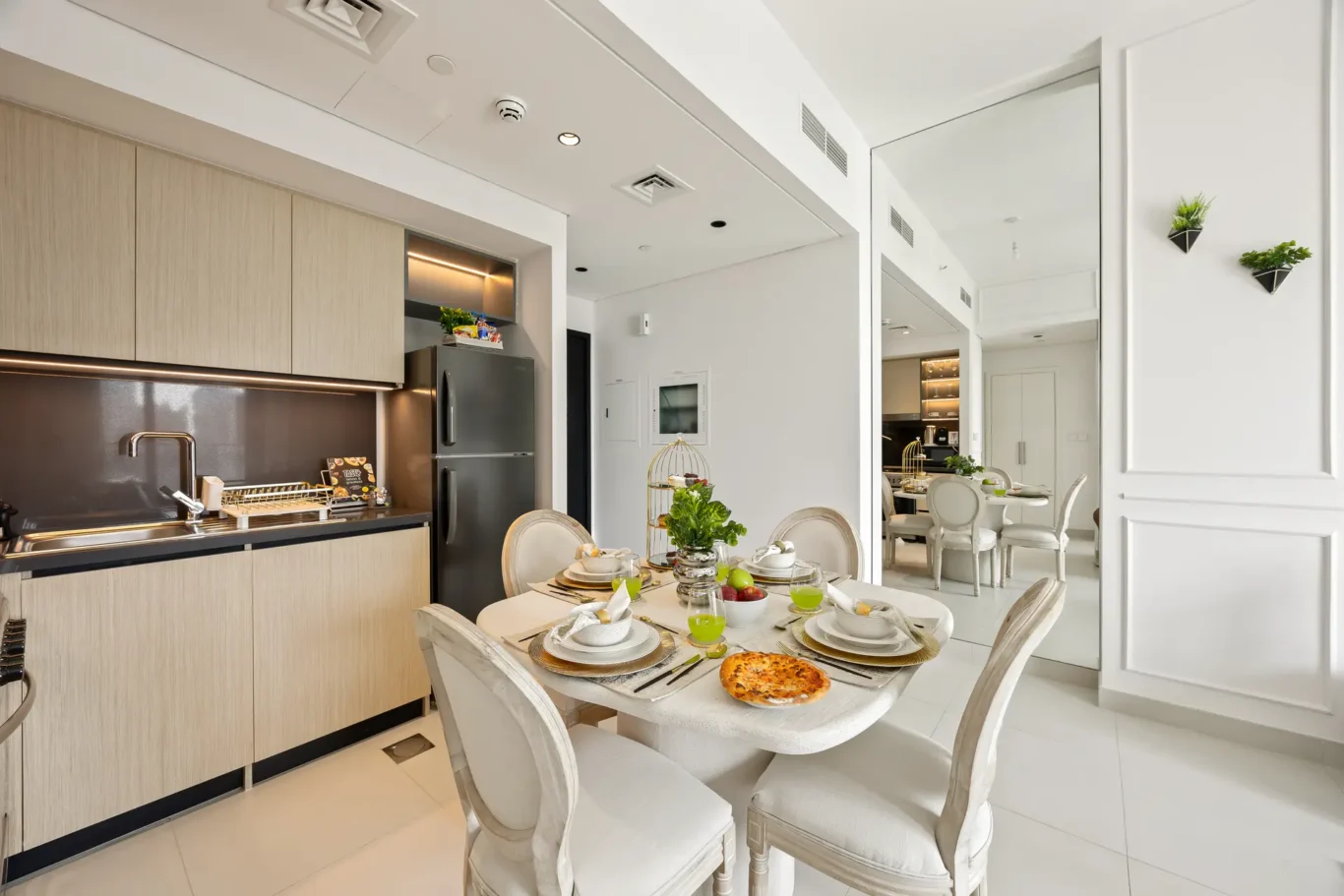 Open-concept kitchen and dining area with light wood cabinets, stainless refrigerator, and a round dining table set for breakfast. A pizza sits on the table under bright lighting.