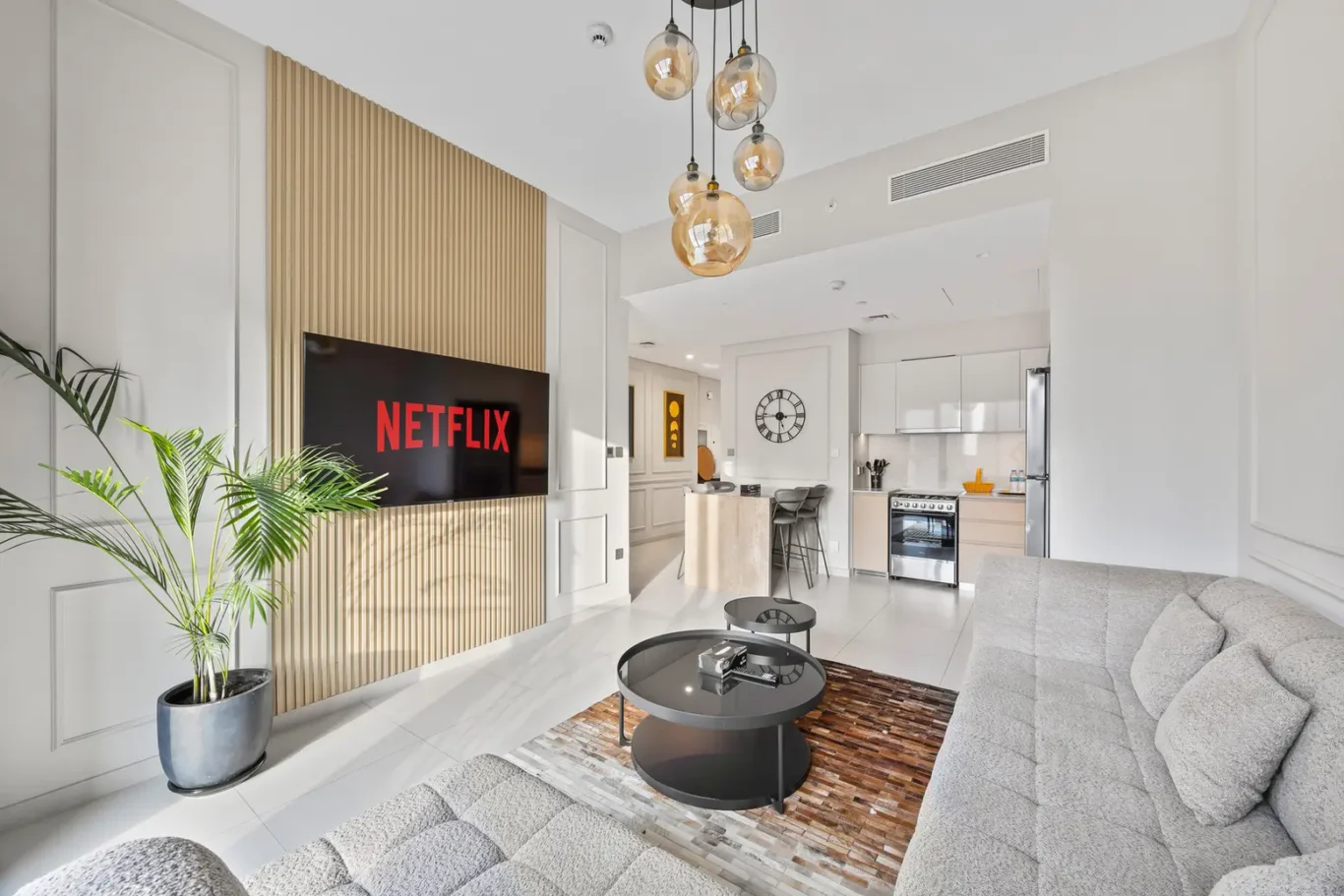 Modern living room with a Netflix screen mounted on a wooden slat accent wall, gray sectional sofa, and glass coffee tables.