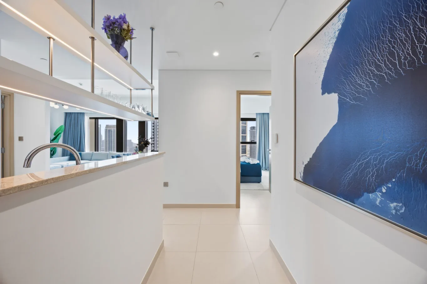 Bright modern hallway with an open kitchen bar on the left, glassware on shelves, and a large blue abstract artwork on the right.