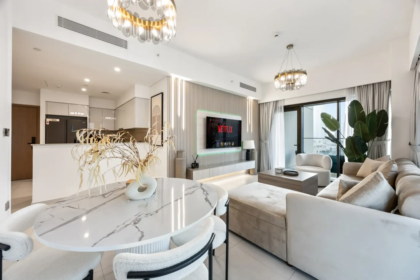Bright open-plan living room and kitchen with a wall-mounted TV showing Netflix, beige sofa, marble dining table, and large windows with curtains.