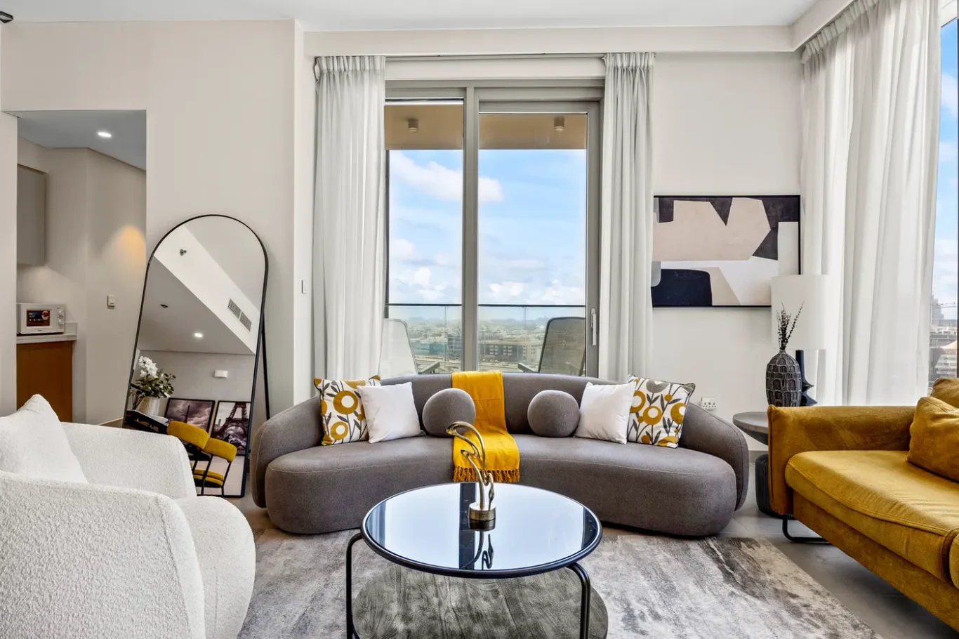 Modern living room with a curved gray sofa, yellow throw, white and patterned cushions, and a round glass coffee table.