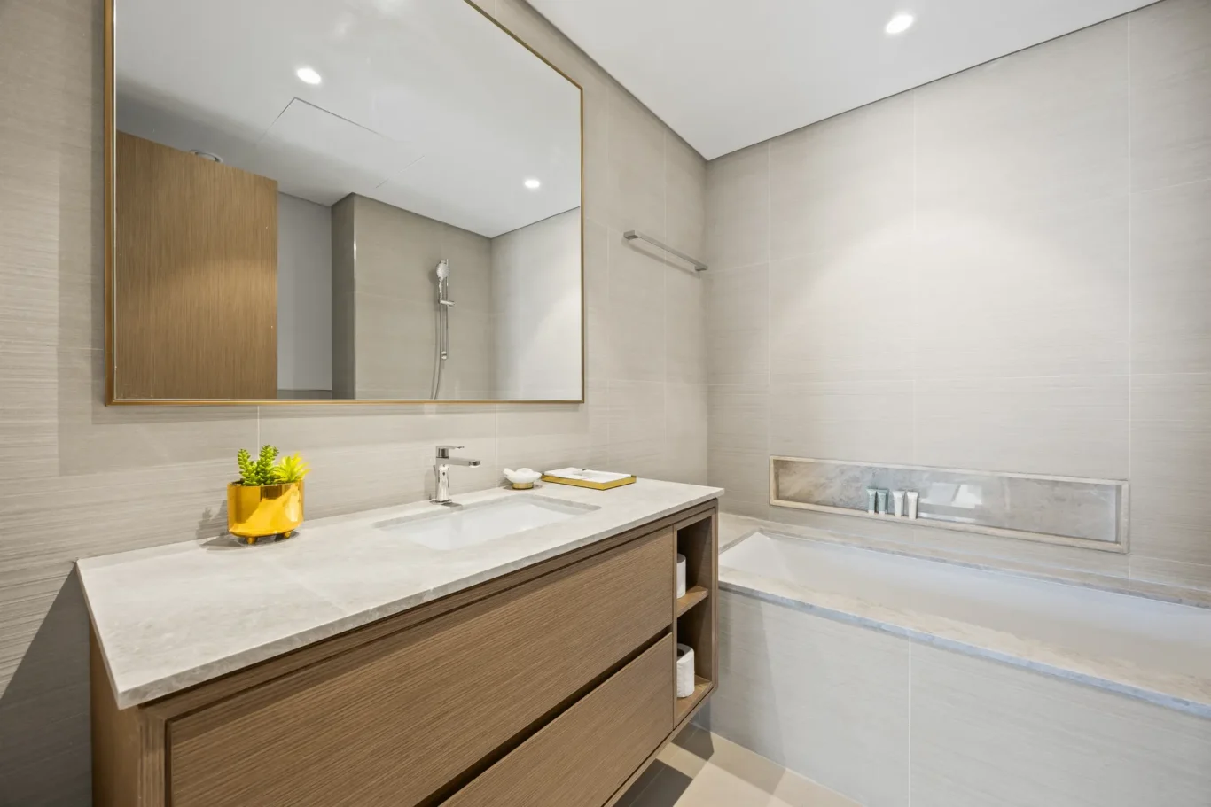 Modern bathroom with a wooden vanity, large gold-framed mirror, and a marble countertop with a small plant in a gold pot. A shower and tub area lines the light-gray tiled wall in the background.