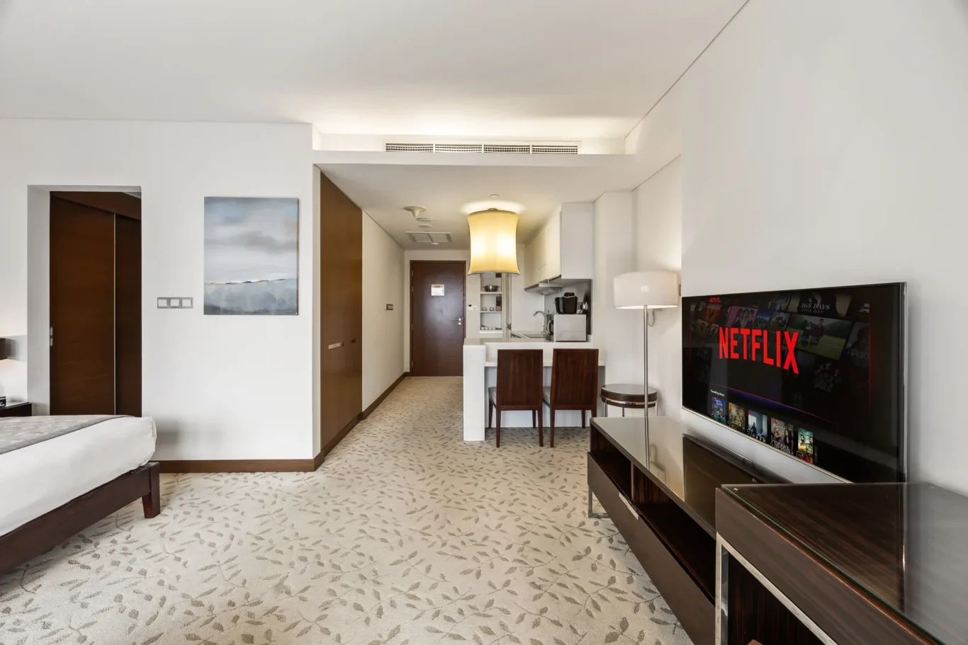 Living area of a hotel suite with a flat-screen Netflix TV, two wooden chairs, a small table, and a kitchenette in the back left-right area.