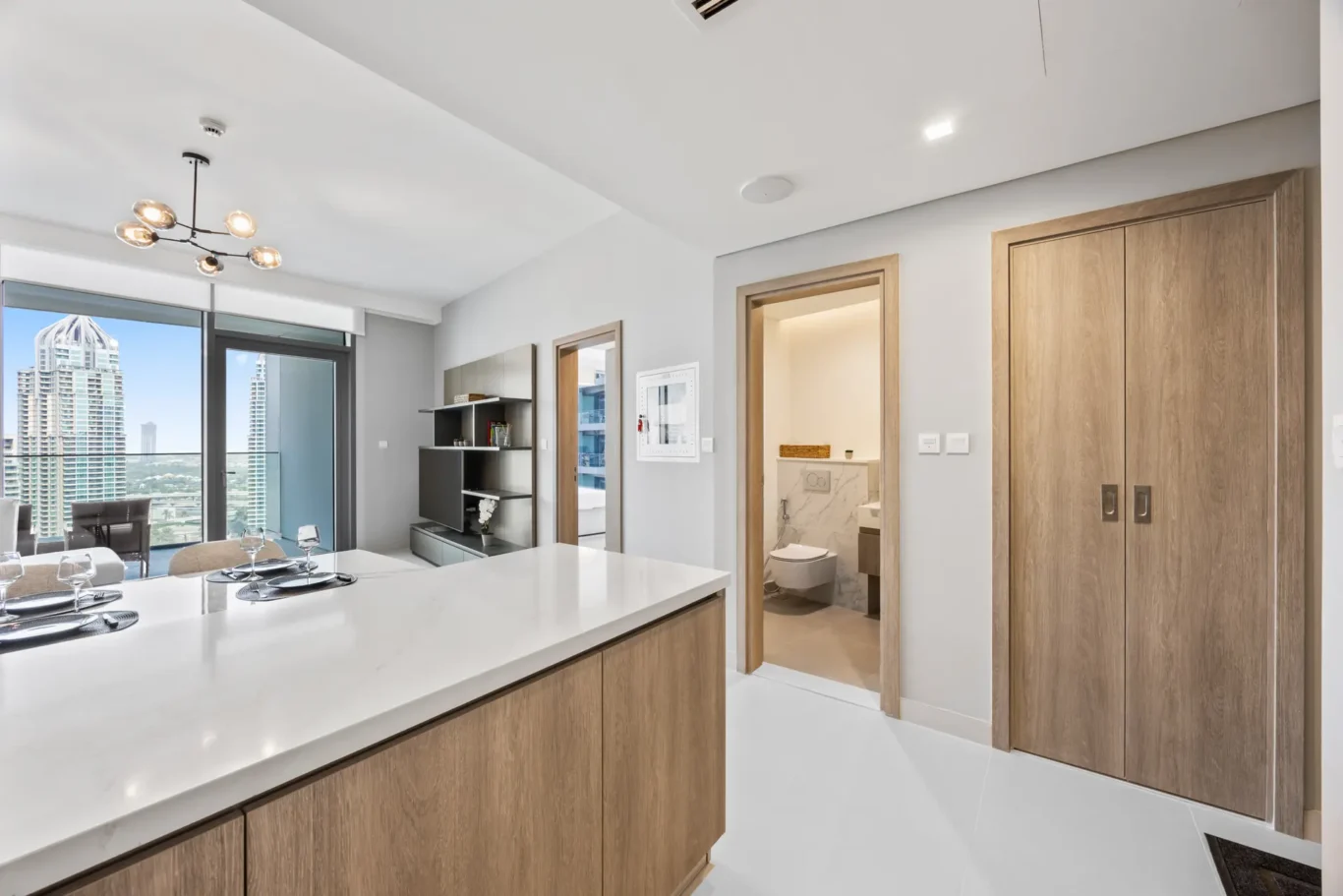 Modern open-plan kitchen with a white island, wooden cabinets, and a dining table; glass doors open to a balcony with city edificios visible outside.