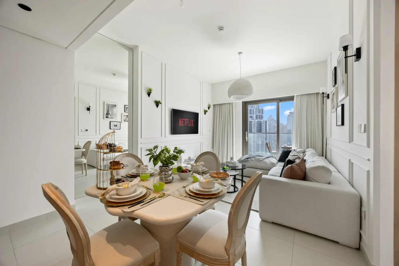 Open-plan dining area with a breakfast spread on a light wood table and a living room with a white sofa, large windows, and a cityscape view.