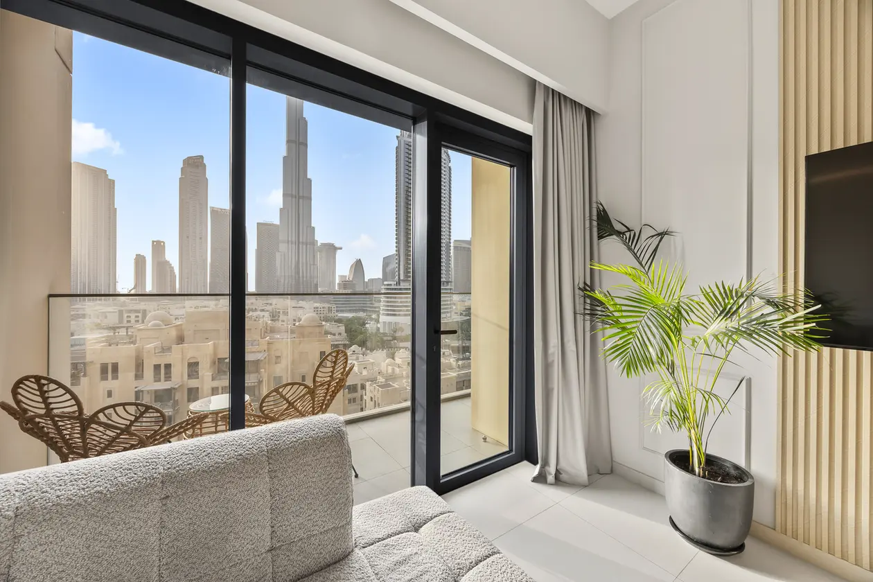 Modern living room with large glass doors opening to a balcony and a city skyline; wicker chairs on the balcony and a tall potted plant inside.