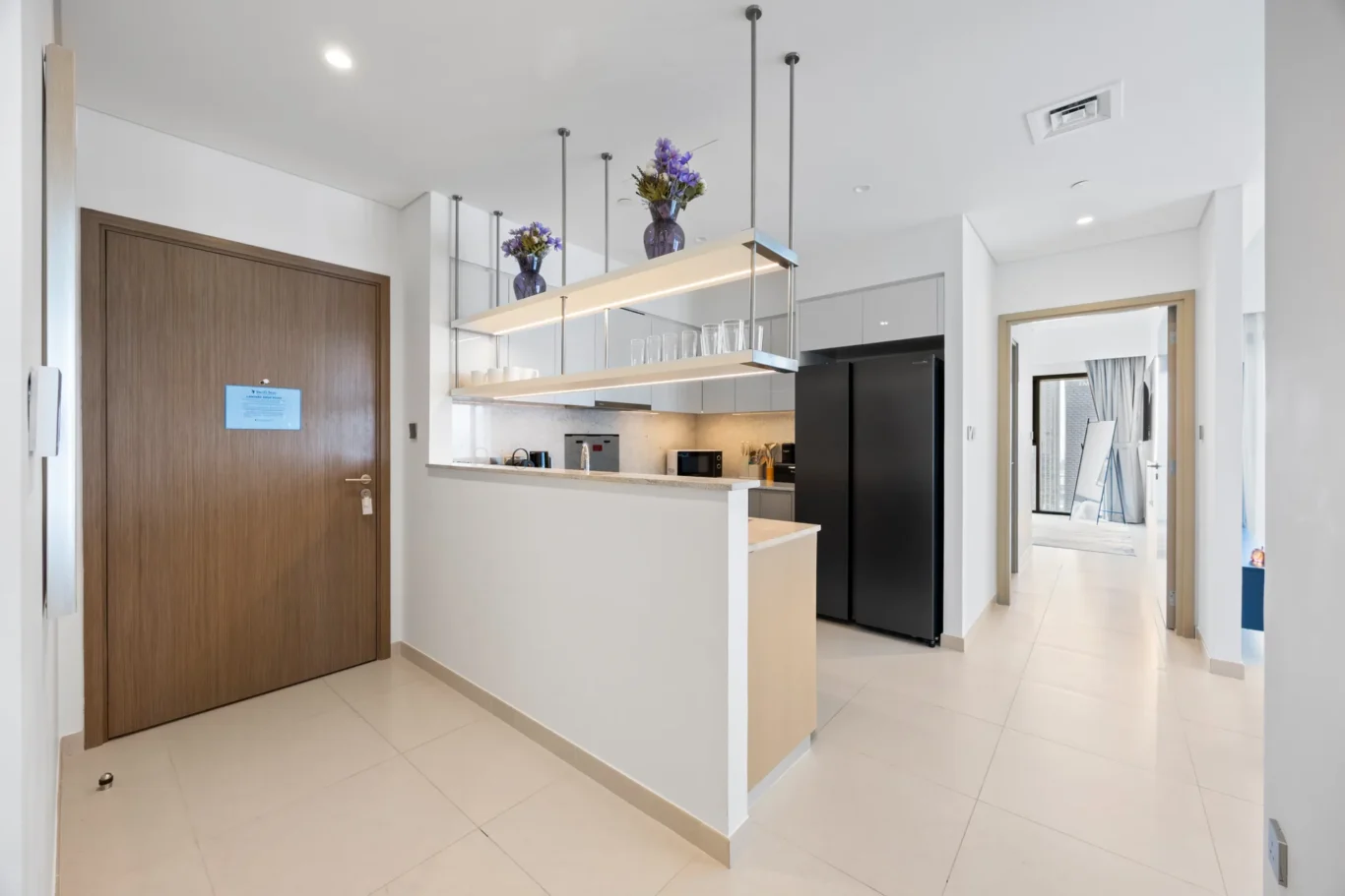 Bright contemporary kitchen with a white island, hanging shelf unit, and a tall black refrigerator visible from a hallway.