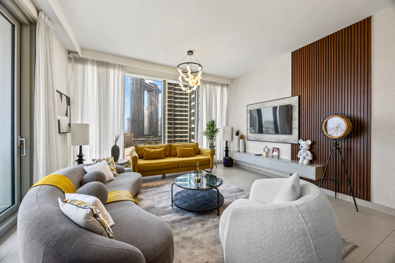 Bright open-concept living room with a mustard sofa, gray curved sofa, white chairs, and a city view through large windows; contemporary decor and coffee table centerpiece.