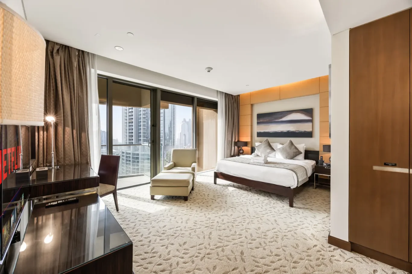 Spacious hotel room with a king bed, seating area, and a balcony overlooking a city skyline through large windows.