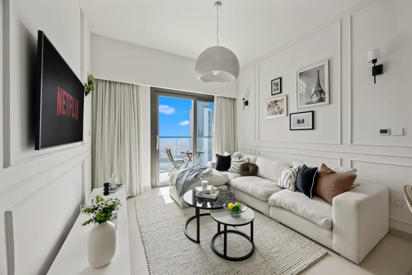 Bright white living room with an L-shaped sofa, round black coffee tables, and a balcony door showing the ocean view